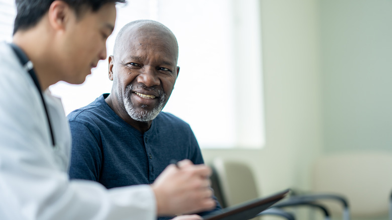 A senior gentleman sits in a waiting room beside his doctor as they review test results on a tablet together.