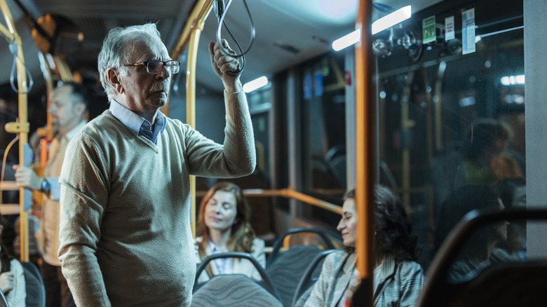 Older man wearing glasses stands on bus while two women sit and other man stands behind him