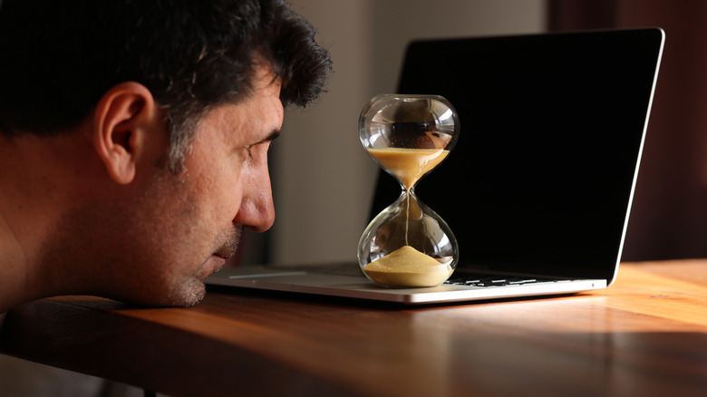 man stares at hour glass sitting on his laptop