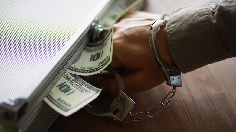 handcuffed hand holding handle of briefcase that has money spilling out