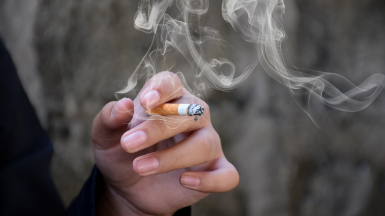 Hand holding a lit cigarette surrounded by smoke