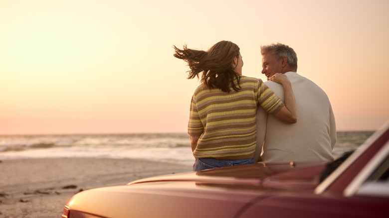 a retired couple on the hood of a car looking at the sunset over the ocean