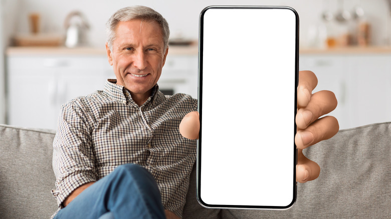 A senior man sitting on a couch while holding up a smartphone screen.