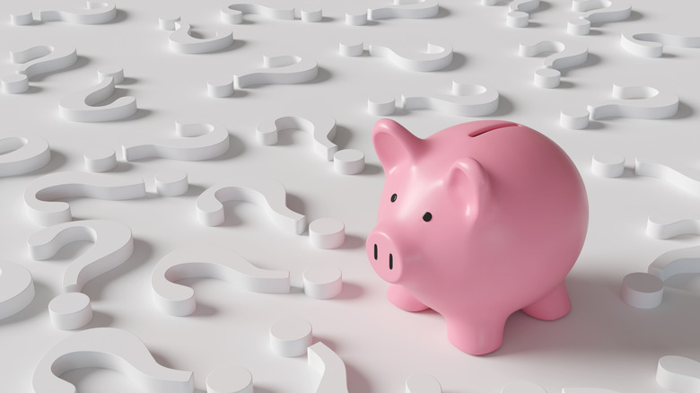 Pink piggy bank standing among white question marks on white background.