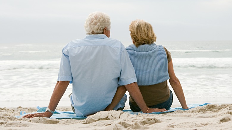 An elderly couple are seated on a blanket at the beach facing the waves