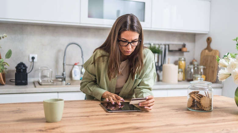 Stressed woman holding credit card and checking tablet in kitchen.