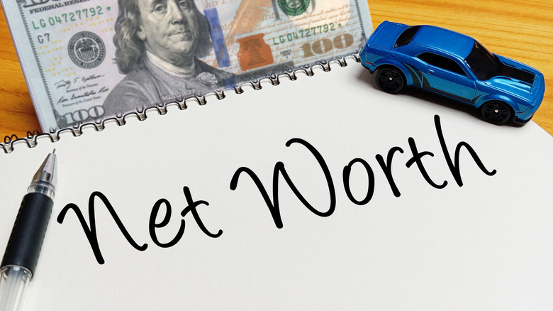 Blue toy car on a spiral notebook with handwritten text Net Worth, a hundred dollar bill, and a pen on a wooden desk.