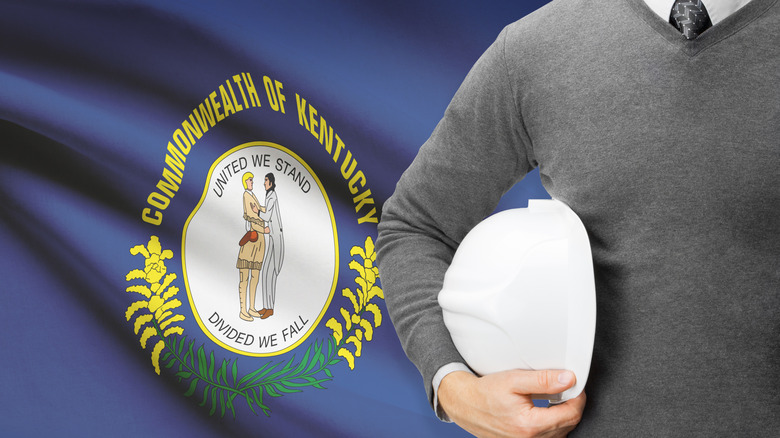 Man in sweater and tie holding hard hat stands in front of Kentucky state flag
