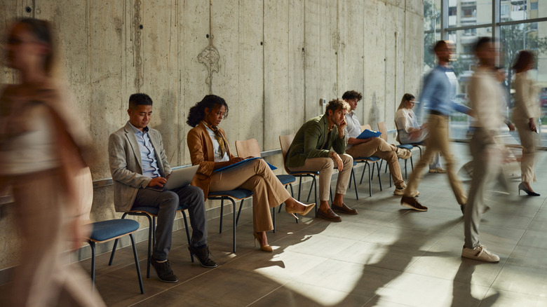 A group of multi-ethnic men and women sit waiting as people walk around