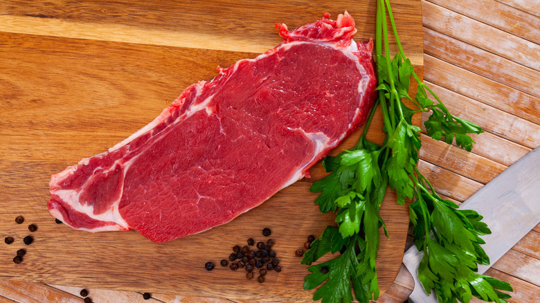 Piece of raw boneless beef on wooden background with seasonings and greens prepared for roasting.