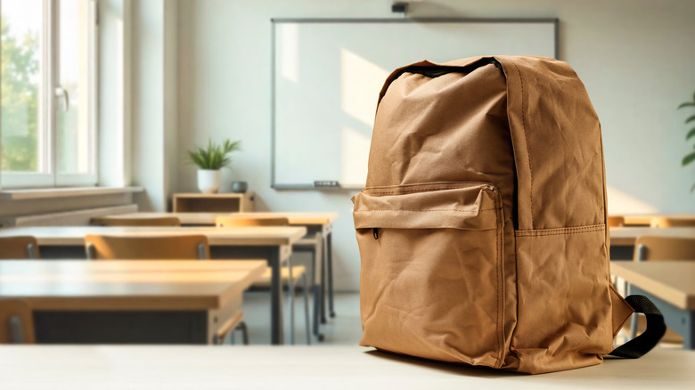 School desk with backpack in foreground and blurred classroom interior in background.