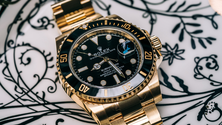 A gold and black Rolex Submariner watch on a white tray with a black floral design.