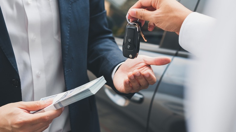 A person hands over car keys to a buyer holding a stack of cash beside a vehicle