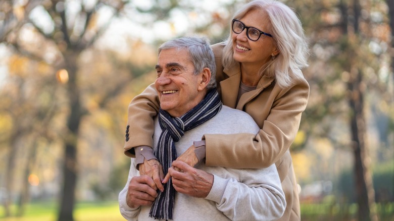 An elderly couple outdoors in a park during autumn, smiling as the woman wraps her arms around the man from behind; trees with fall foliage blur in the background