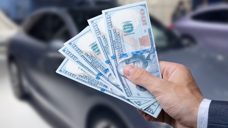 Hand holding several U.S. $100 bills in front of a parked car