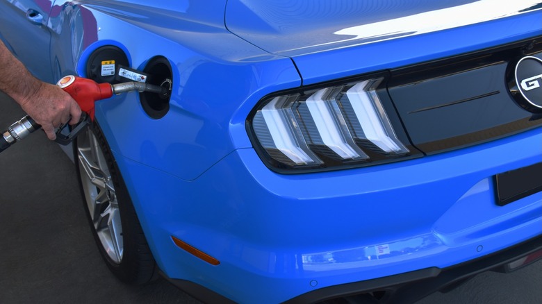 Close-up of a blue Ford Mustang being refueled at a gas station, showing the rear taillights and fuel nozzle inserted into the fuel port