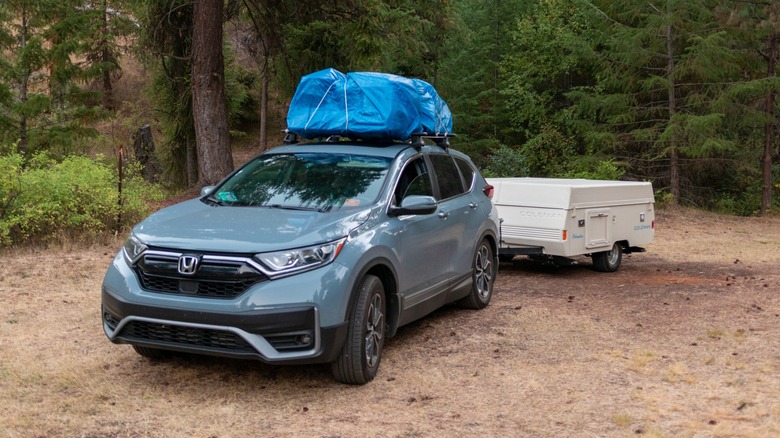 Honda CR-V parked at a forest campsite with a blue cargo bag on the roof and a small pop-up camper trailer attached