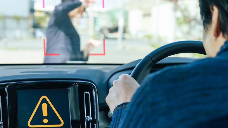 Driver's-eye view inside a car as forward collision warning highlights a pedestrian crossing the road, with a dashboard alert symbol visible on the center display