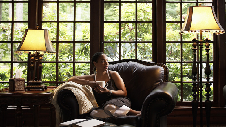 A middle-aged woman relaxing on a sofa in her home in front of expansive windows.