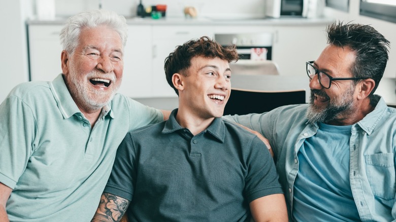 A younger man laughing and sitting between a baby boomer and a middle-aged man on a couch