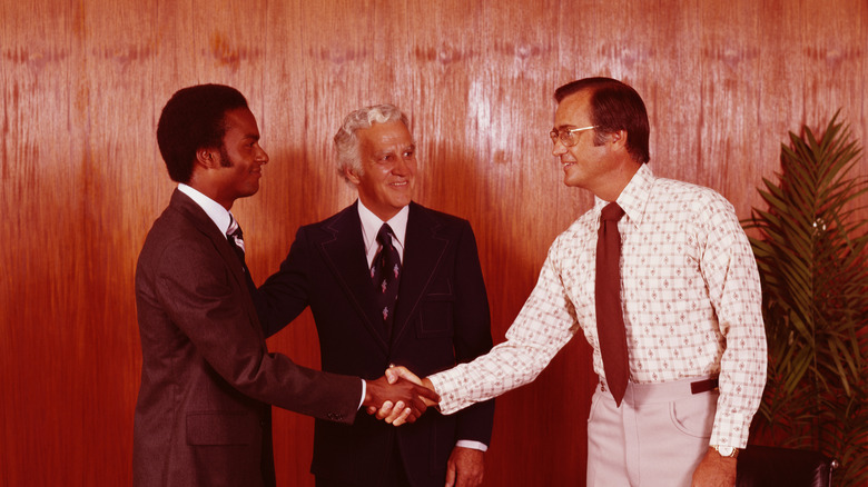 two men in 1970s business attire in an office being introduced by a third man