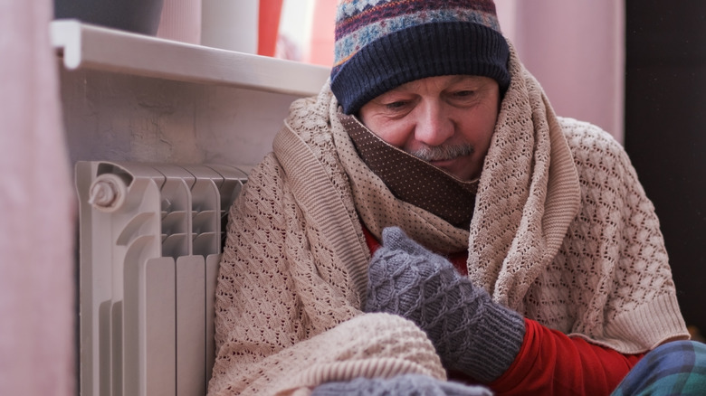 Man in cold weather clothes and blankets huddles next to a heater