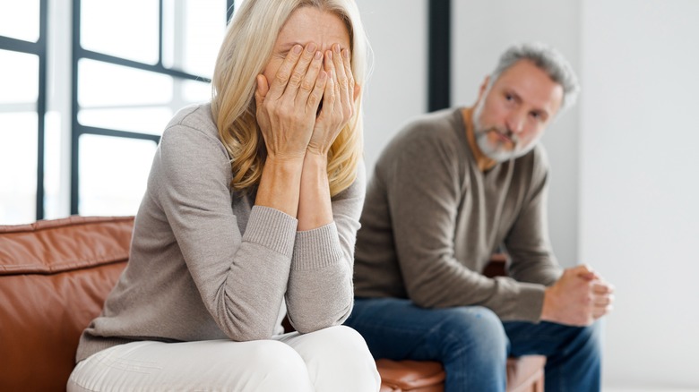 Woman on couch covers face while man seated next to her stares