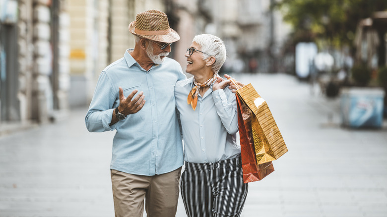 A smiling retired couple walking with their arms around each other and the female holding shopping bags.