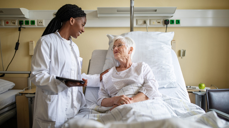 A smiling young nurse places a hand on the shoulder of a smiling but bed ridden senior woman in a hospital.