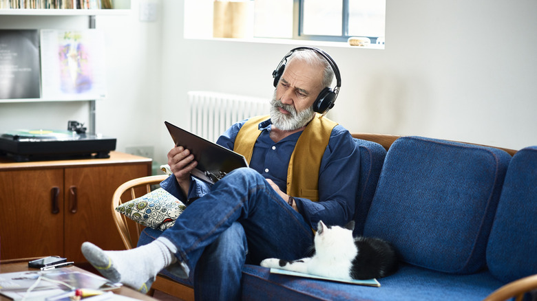 A senior man wearing headphones while sitting on a couch next to a cat