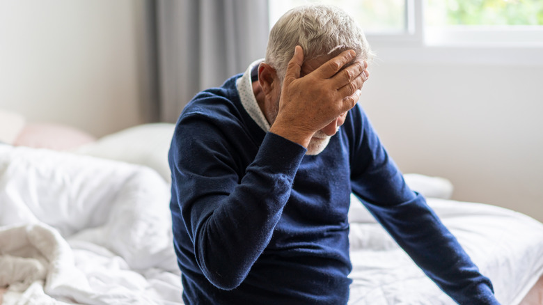 A distraught elderly man sitting in bed with a hand over his face.
