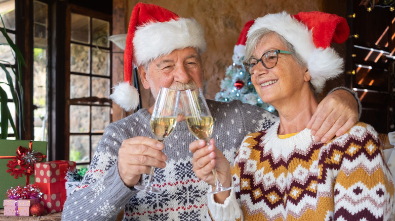 An older couple in Santa hats and Christmas sweaters clink glasses of champagne