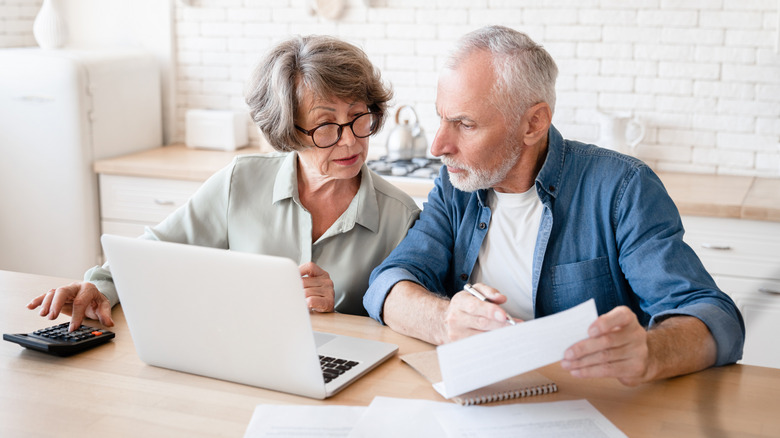 An older couple look worried while making calculations at a table, computer open in front of them.