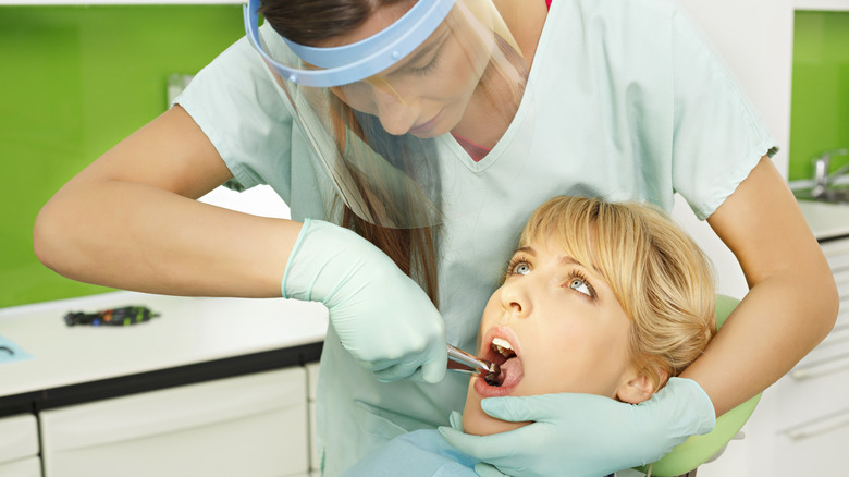 white female dentist wearing face shield extracts tooth from white female patient