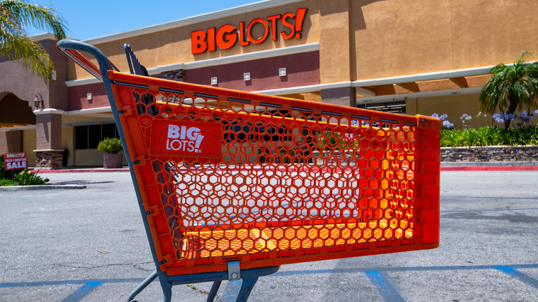 A Big Lots storefront and parking lot on a sunny day, with a bright orange Big Lots! shopping cart in the foreground.
