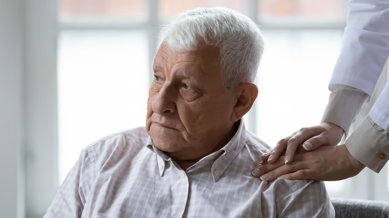 An older man looking upset, with the hands of a caregiver on his shoulder.