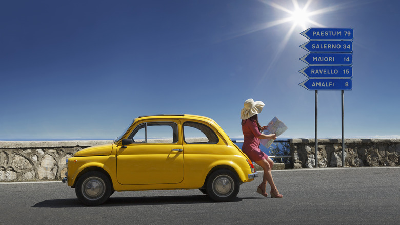Young caucasian woman leaning on her vintage Fiat 500 car, checking her map on the road along the Amalfi Coast.