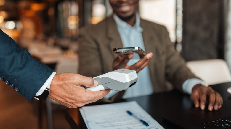 A hand offers a payment device to the man as he holds his phone.