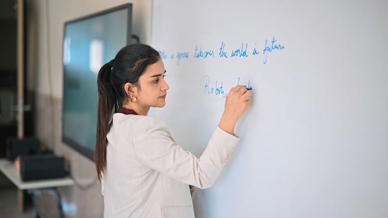 A teacher in a light blazer neatly writes a lesson topic on the whiteboard.