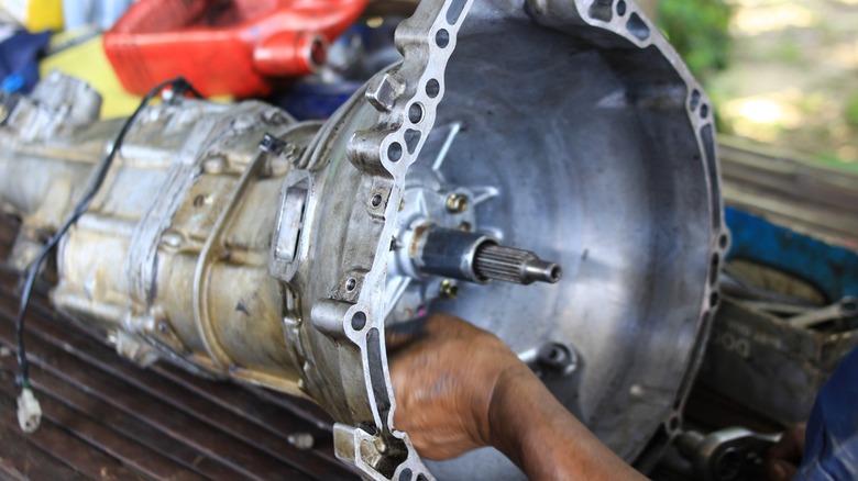 Close-up of a removed automatic transmission on a workbench, showing the bell housing and input shaft while a mechanic works on it in the background