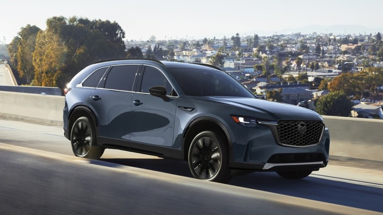 bluish Mazda CX-90 moving on highway above residential are in background