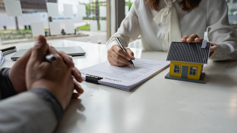Faceless woman fills out a contract while touching a model house as another faceless man with hands surrounding a pen sits opposite her from a table.