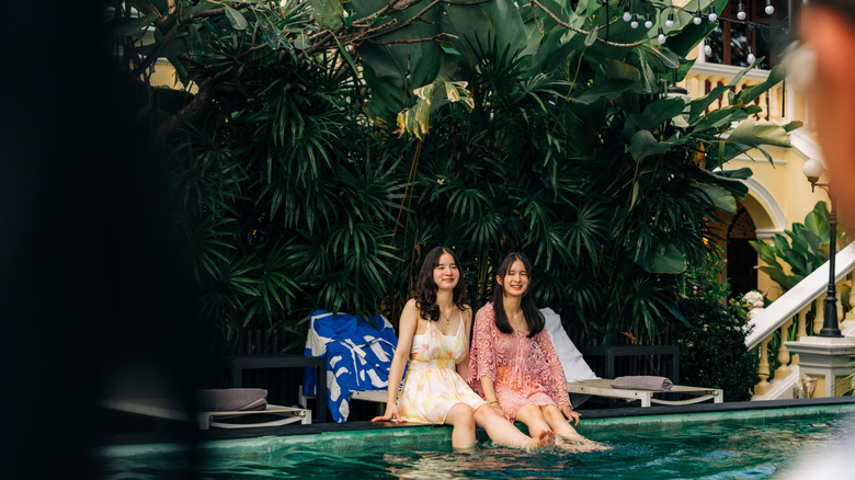 Two young women sitting by side of pool in villa.