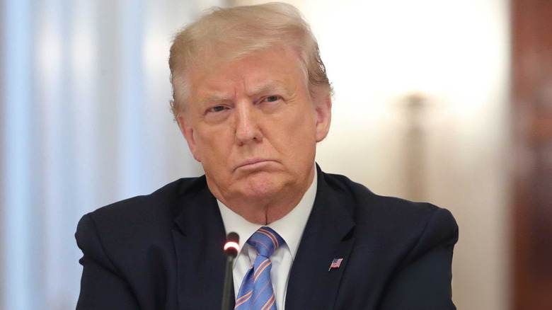 President Trump frowns while wearing an American flag lapel pin, in front of a blurred white background.