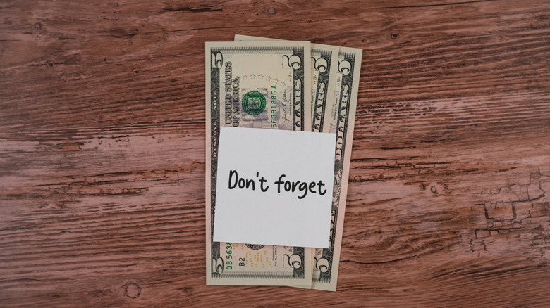 Three five-dollar bills laid out on a wooden tabletop with a sticky note that reads: "Don't forget."