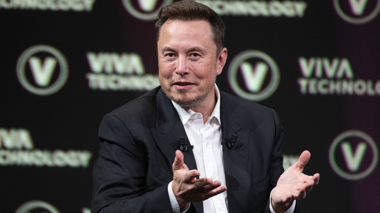 Elon Musk, in black suit, raises hands up in questioning, supplicant gesture in front of a Viva Technology step-and-repeat.