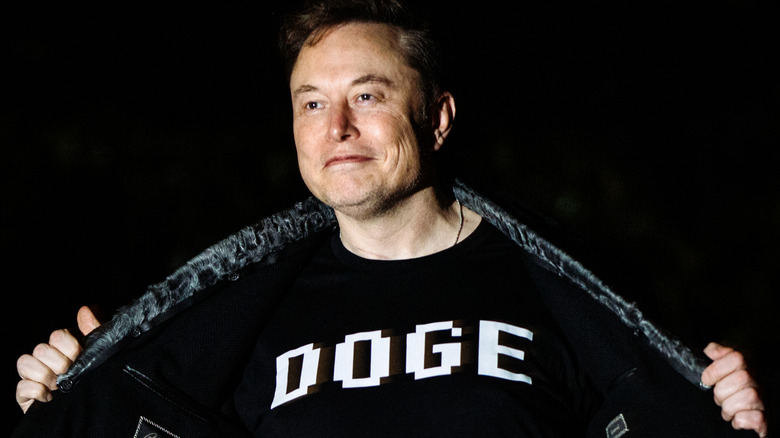 A grinning Elon Musk opening his jacket to reveal a DOGE t-shirt