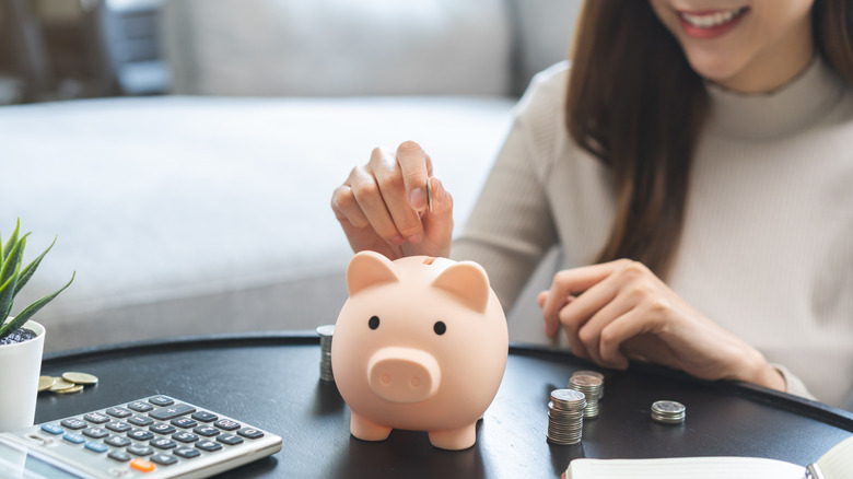 Smiling woman puts coin in piggie bank while sitting at tale with coins and calculator