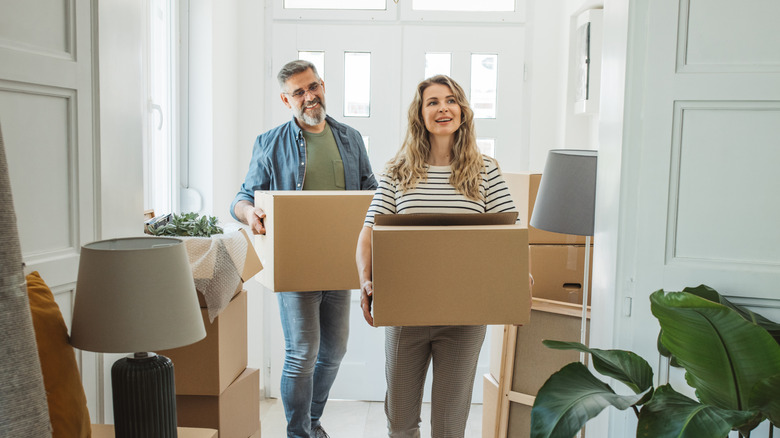 Smiling man and woman each holding a box as they walk into a room filled with boxes and furniture