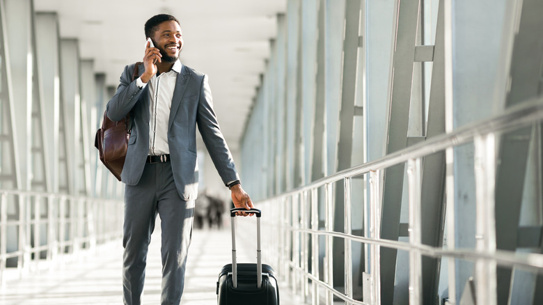 Business man on phone pulling luggage through airport